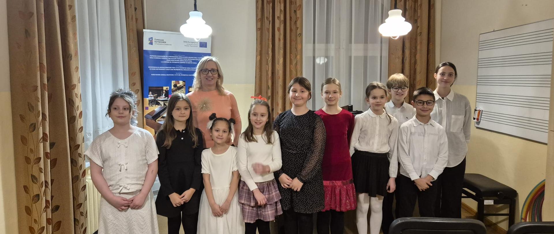 Na zdjęciu widać grupę kilkunastu osób pozujących do wspólnego zdjęcia w sali muzycznej. W środku stoi dorosła kobieta (prawdopodobnie nauczycielka lub opiekunka), w jasnoróżowej bluzce, w okularach, z jasnymi włosami do ramion. Uśmiecha się i patrzy w stronę aparatu. Po obu stronach kobiety stoi około 12 dzieci, mniej więcej w wieku szkolnym. Dzieci ustawione są w jednym rzędzie, blisko siebie, wszystkie zwrócone przodem do aparatu. Większość dzieci ma eleganckie, odświętne ubrania: dziewczynki w sukienkach lub spódnicach (białe, czarne, bordowe, wzorzyste), chłopcy w białych koszulach i ciemnych spodniach. Dzieci mają różne fryzury; część dziewczynek ma włosy rozpuszczone lub związane, jedno dziecko ma we włosach czerwoną kokardkę. Wszyscy wyglądają spokojnie i skupieni, niektórzy lekko się uśmiechają. Pomieszczenie przypomina salę do nauki muzyki. Po prawej stronie ściany widać tablicę z pięciolinią muzyczną. Z tyłu stoją okna z jasnymi firankami i brązowymi zasłonami. Z sufitu zwisają lampy na długich czarnych przewodach, świecące ciepłym światłem. Po lewej stronie w tle znajduje się roll-up lub plakat informacyjny. Na pierwszym planie, u dołu zdjęcia, widać kilka czarnych krzeseł, ustawionych tyłem do aparatu, jak dla publiczności.