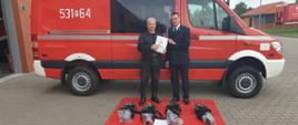 Komendant Powiatowy PSP wraz z Prezesem OSP, w tle samochód strażacki, na ziemi 4 komplety aparatów powietrznych.