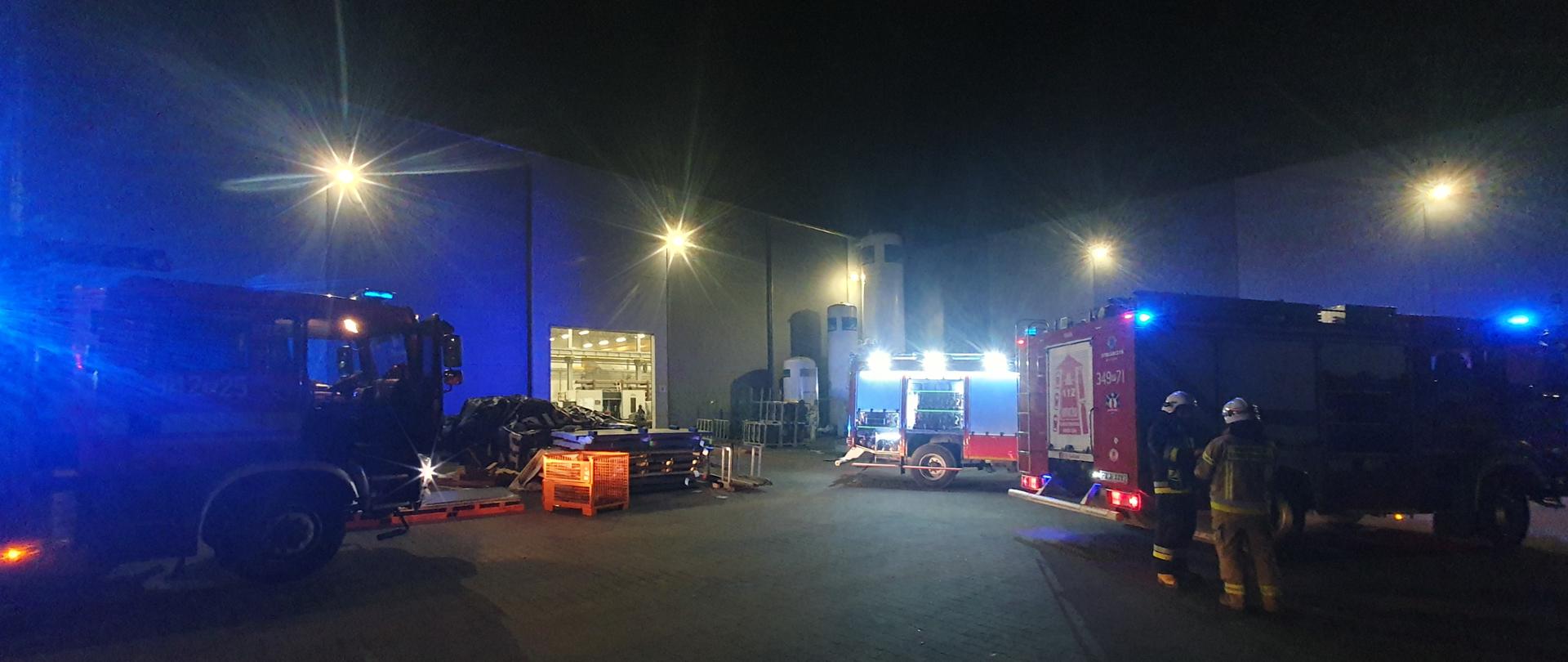 Zdjęcie hali w porze nocnej. Przed halą pojazdy ratowniczo-gaśnicze z włączoną sygnalizacją ostrzegawczą. Sztuczne oświetlenie terenu działań. Widać ratowników.