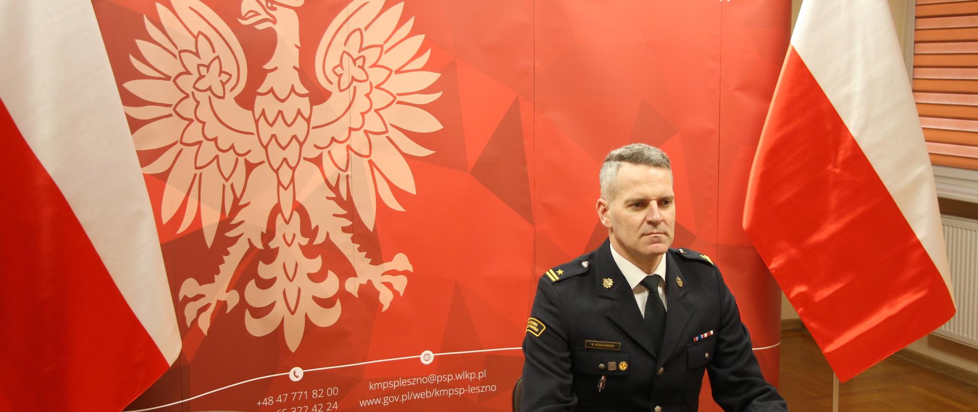 Osoba w mundurze siedzi przy biurku w tle widoczne flabi i godło.