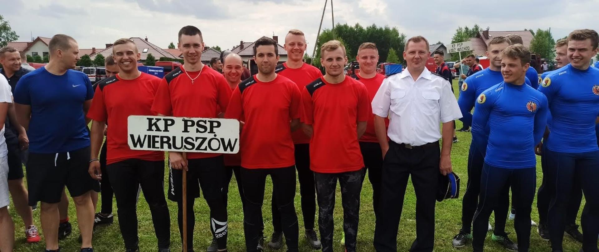 Zdjęcie przedstawia strażaków z Komendy Powiatowej Państwowej straży Pożarnej w Wieruszowie stojących na boisku sportowym. Jeden ze strażaków trzyma w ręku transparent z nazwą jednostki reprezentującej w zawodach.