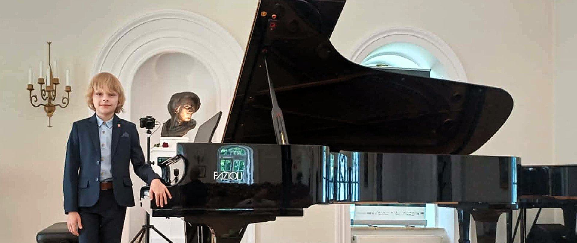 chłopiec ubrany na galowo stojący przy otwartym fortepianie we wnętrzu zabytkowej sali, po lewej fragment drugiego fortepianu a zanim stojący baner