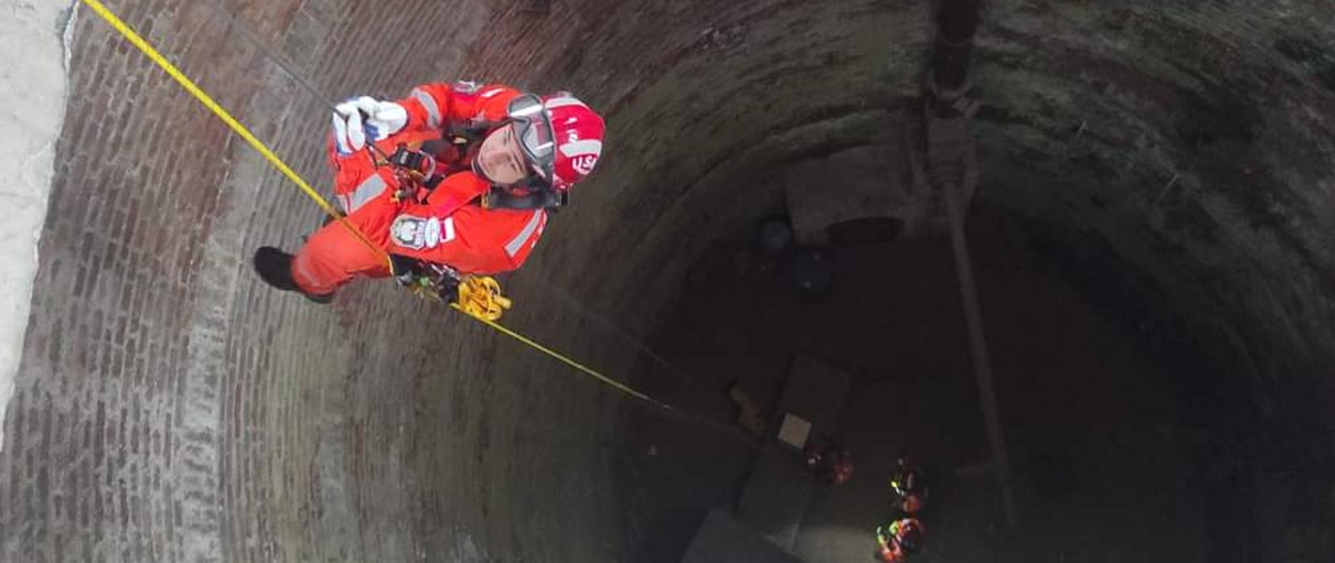Funkcjonariusz Państwowej Straży Pożarnej podczas ćwiczeń. Podpięty pod linki, w asekuracji opuszcza się w dół.