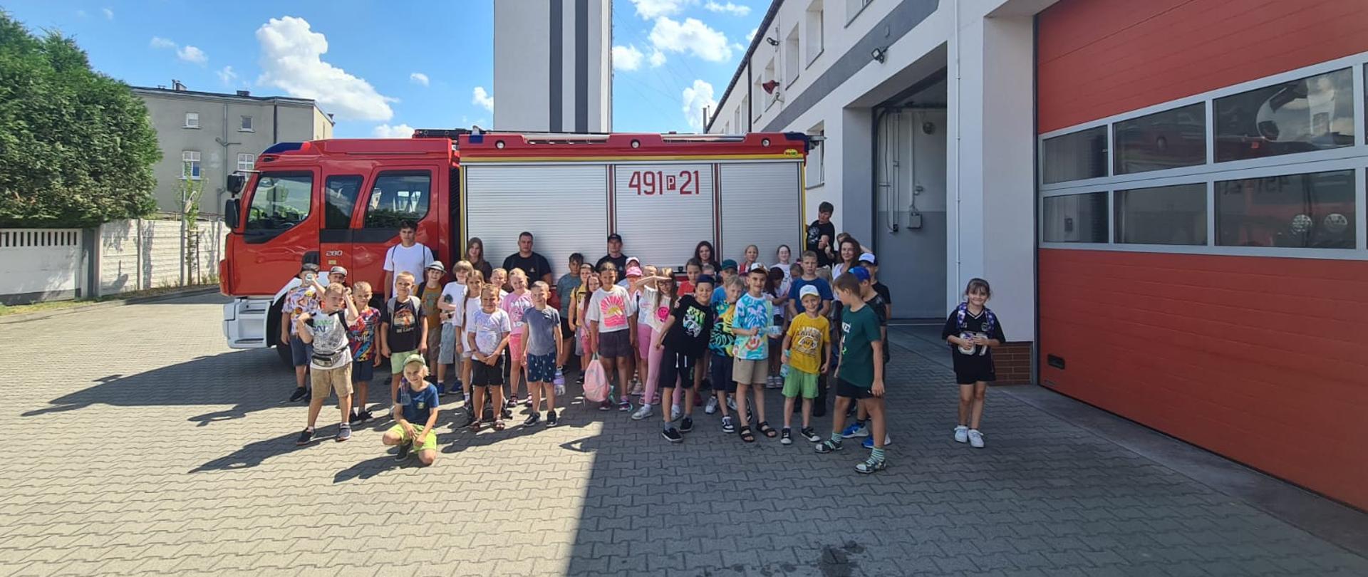 Grupa dzieci i dorosłych pozuje przed czerwonym wozem strażackim na tle remizy strażackiej w słoneczny dzień