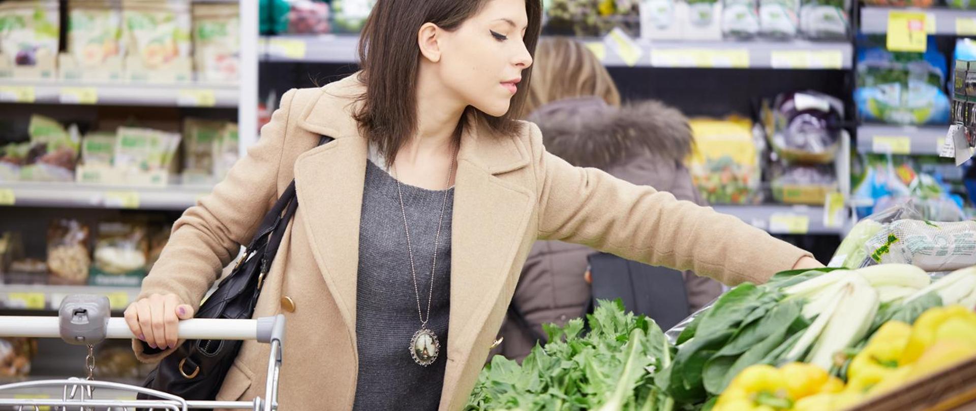 Kobieta przy stoisku warzywnym wybiera produkty do kosza zakupowego.