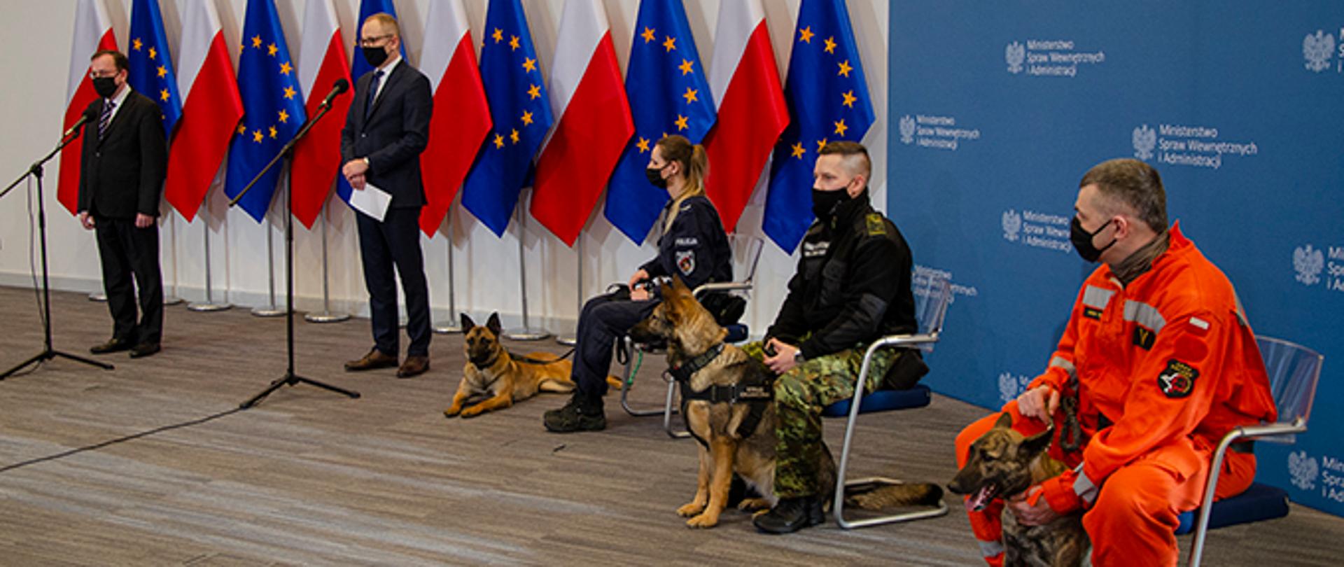widok sali konferencyjnej, przedstawiciele MSWiA na tle flag Polski oraz Unii Europejskiej, po prawej stronie na krzesłach troje funkcjonariuszy w mundurach z psami