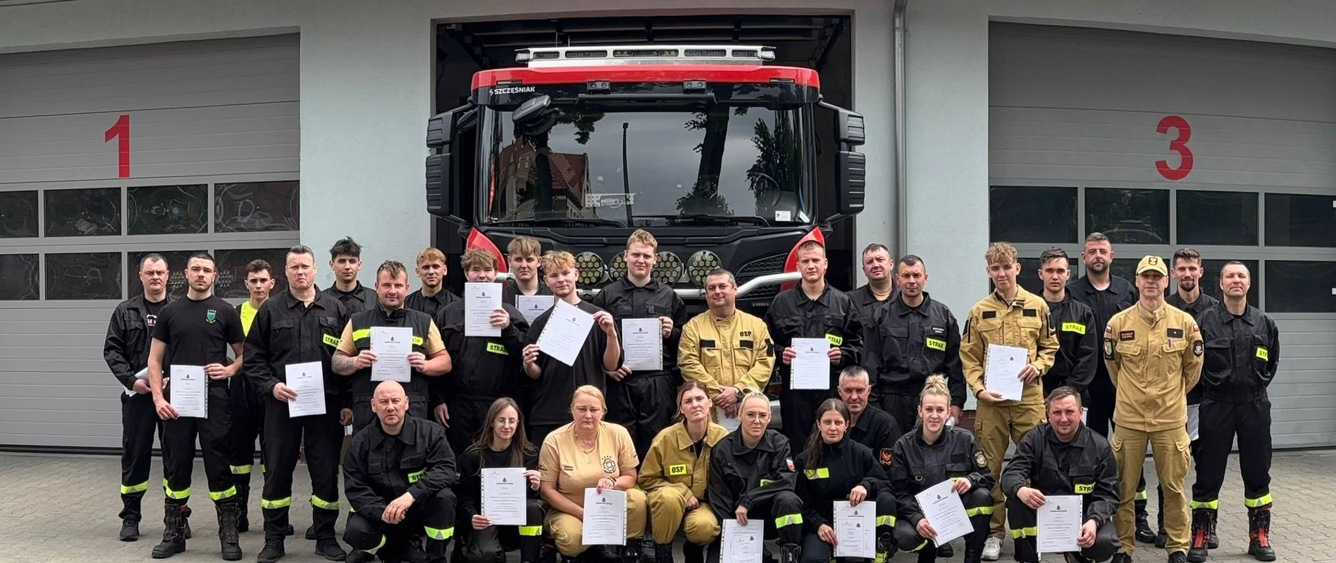 Uczestnicy kursu z dyplomami na tle samochodu gaśniczego i garaży JRG