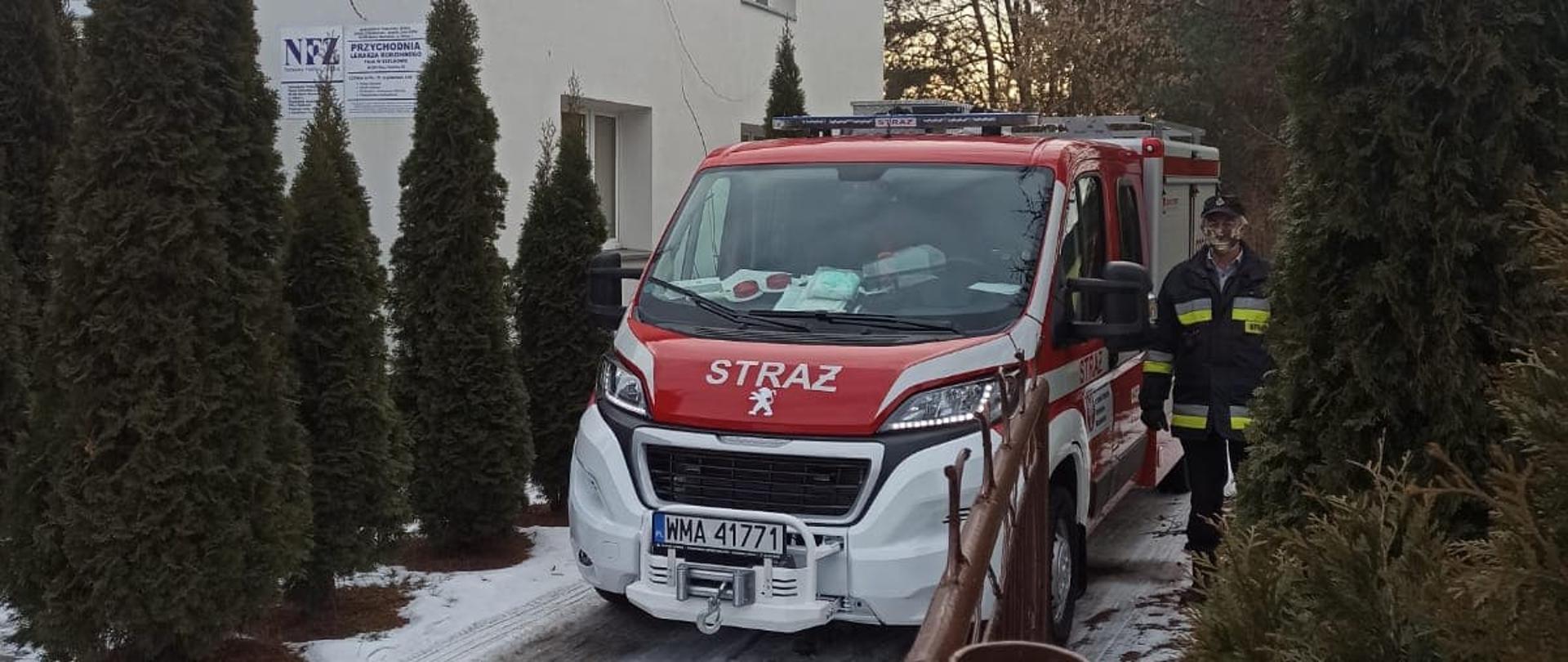 Samochód strażacki OSP Szelków stoi zaparkowany na podjeździe przed przychodnią lekarską. Obok samochodu po jego lewej stronie stoi kierowca.