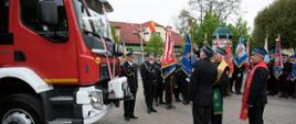 Widok z tyłu. Kapelan małopolskich strażaków w asyście drugiego kapłana dokonuje aktu poświęcenia samochodu pożarniczego, który stoi po lewej stronie. W tle widoczne poczty sztandarowe OSP.