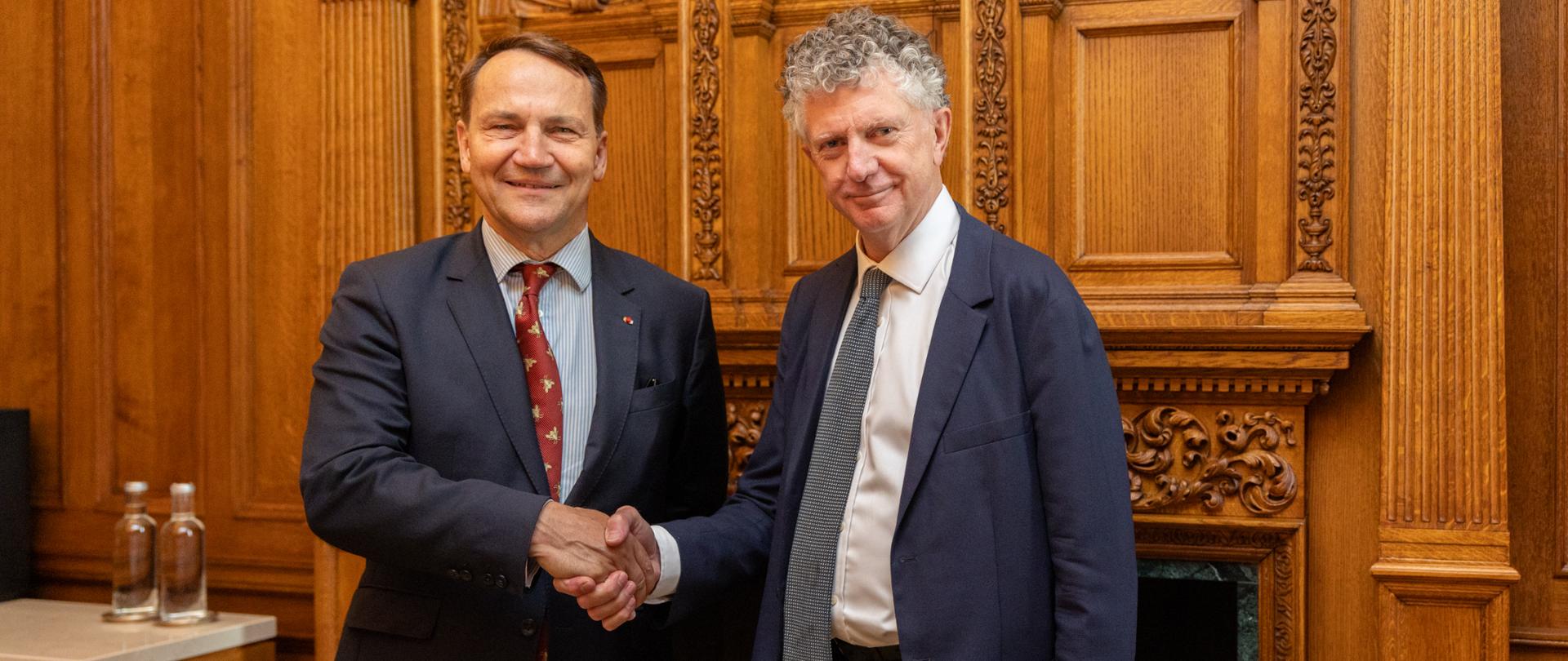 Deputy Prime Minister, Minister of Foreign Affairs of Poland and UK's National Security Adviser, Jonathan Powell in formal business suits shake hands during an official meeting. The meeting takes place in an elegant interior featuring ornate wooden paneling and a fireplace in the background. A small table with two bottles of water is visible on the left side of the room.