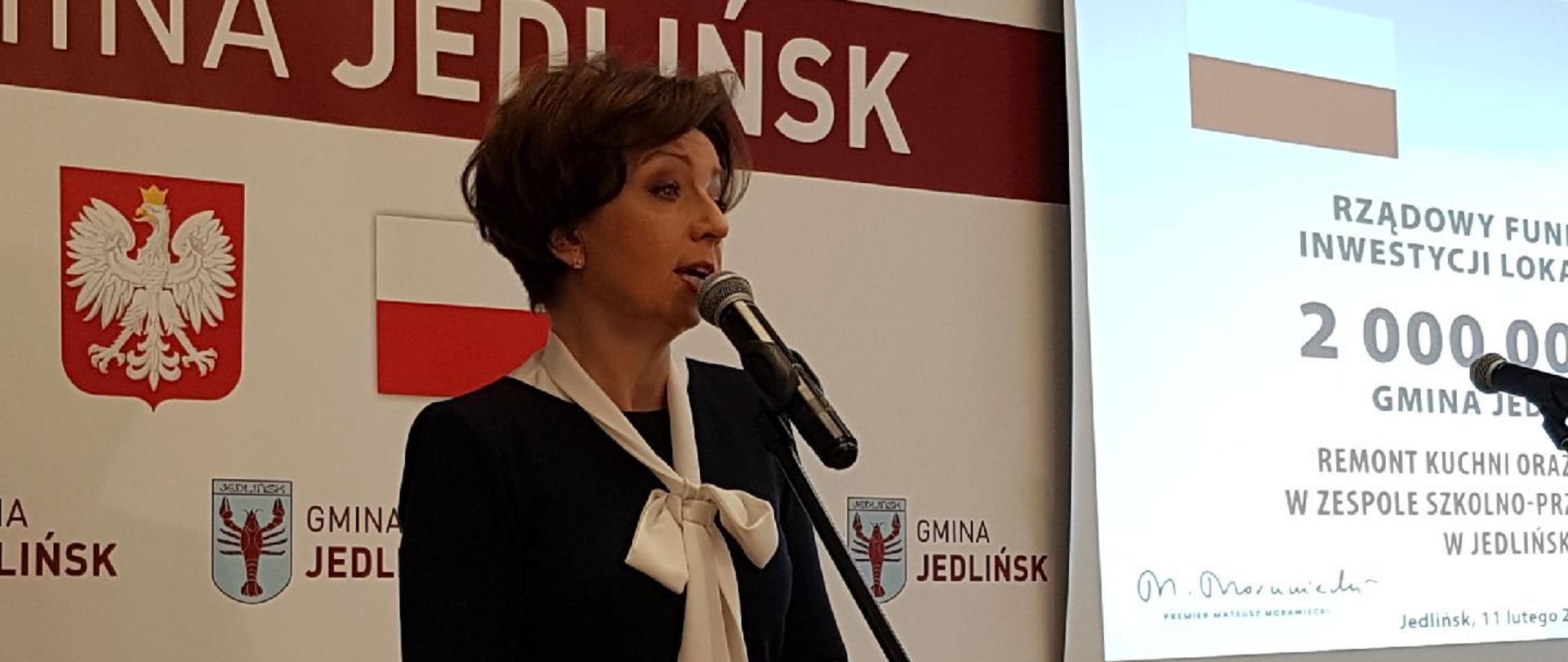 Na zdjęciu przemawiająca minister Maląg. W tle biały baner z godłem Polski, flagą państwową oraz herbem gminy Jedlińsk.