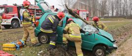 Samochód osobowy w rowie przygnieciony słupem elektrycznym. Wokół strażacy prowadzący działania ratownicze. W tle dwa wozy bojowe PSP