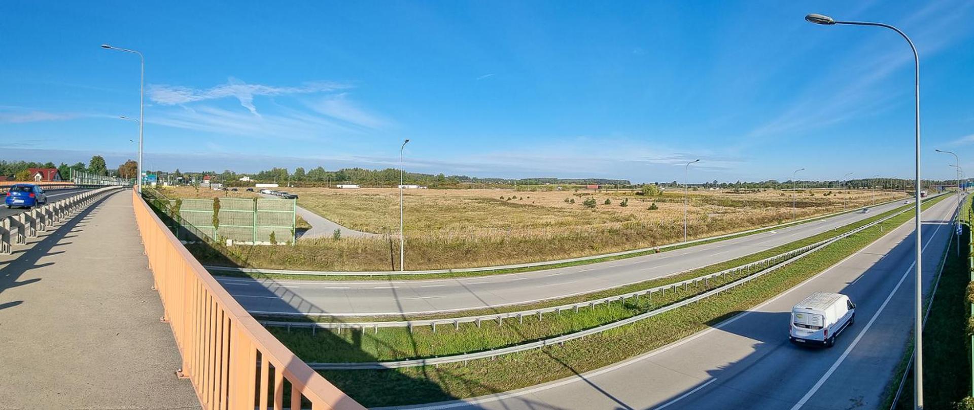 Panoramiczne ujęcie przedstawia rozległy fragment infrastruktury drogowej. Na pierwszym planie widoczna jest estakada z pomarańczowymi barierkami i latarniami ulicznymi, przebiegająca nad jezdnią. Po lewej stronie znajduje się chodnik dla pieszych, wzdłuż którego jedzie niebieski samochód. Pod estakadą, na prawym pasie drogi, porusza się biały van, a pasy ruchu oddziela zielony pas trawy. W tle rozciągają się otwarte tereny z pojedynczymi drzewami i budynkami, a całość znajduje się pod bezchmurnym, błękitnym niebem.