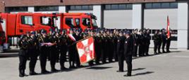 Na pierwszym planie widoczni strażacy biorący udział w uroczystości, za nimi ustawione samochody gaśnicze. W tle budynek komendy. 