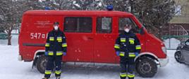 Strażacy wspierają Narodowy Program Szczepień. Na zdjęciu dwóch strażaków w mundurze bojowym stoi na tle samochodu strażackiego, busa. Pojazd stoi na zaśnieżonej drodze. W tle budynek przychodni.
