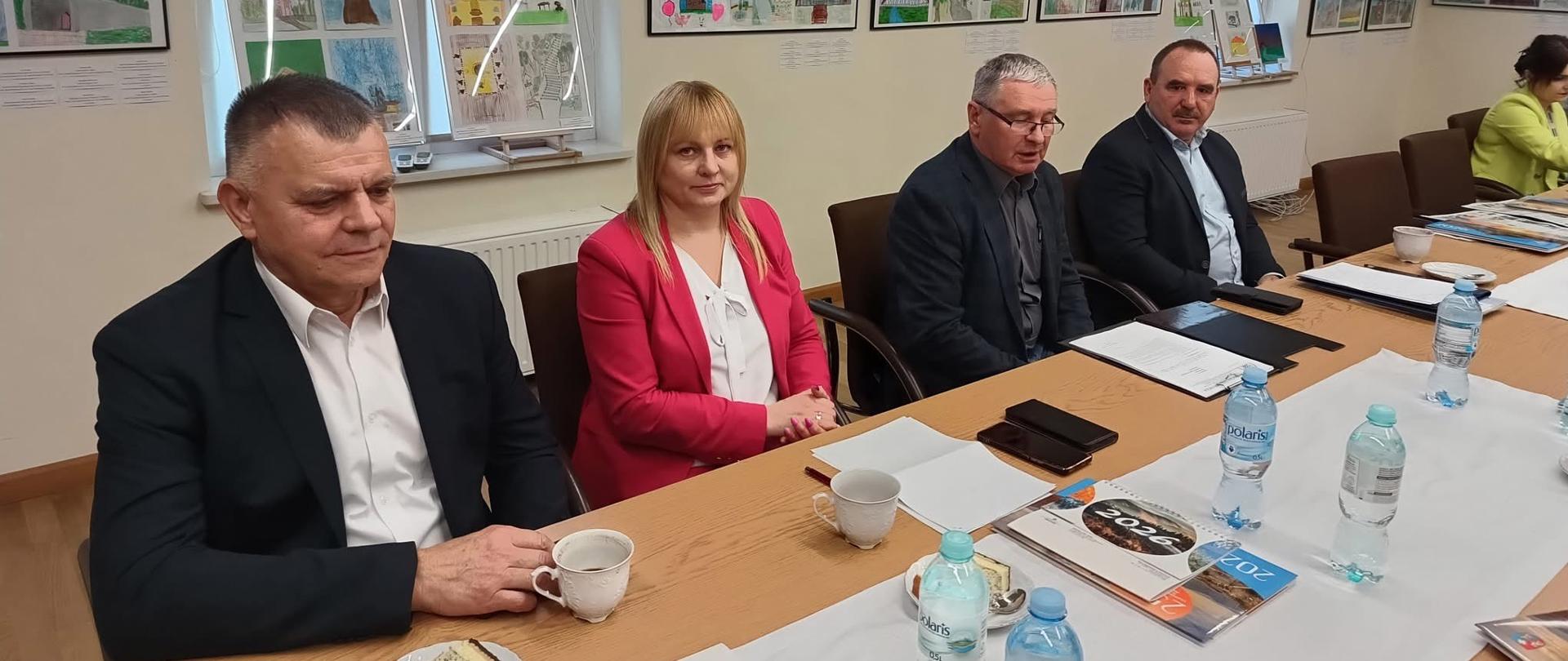 Grupa osób siedząca przy stole. Znajdują się w jasnym pomieszczeniu. Na ścianach wisi dużo kolorowych obrazów.