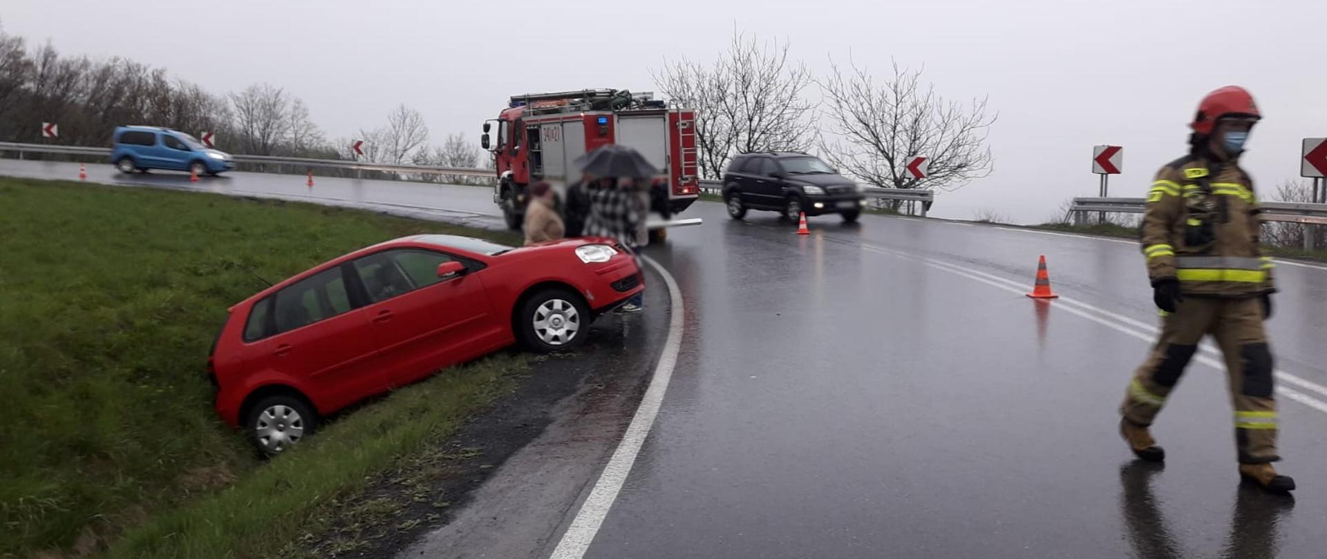 Samochód osobowy koloru czerwonego w rowie. W tle stoi samochód pożarniczy. Drogą idzie strażak oraz jadą samochody. 