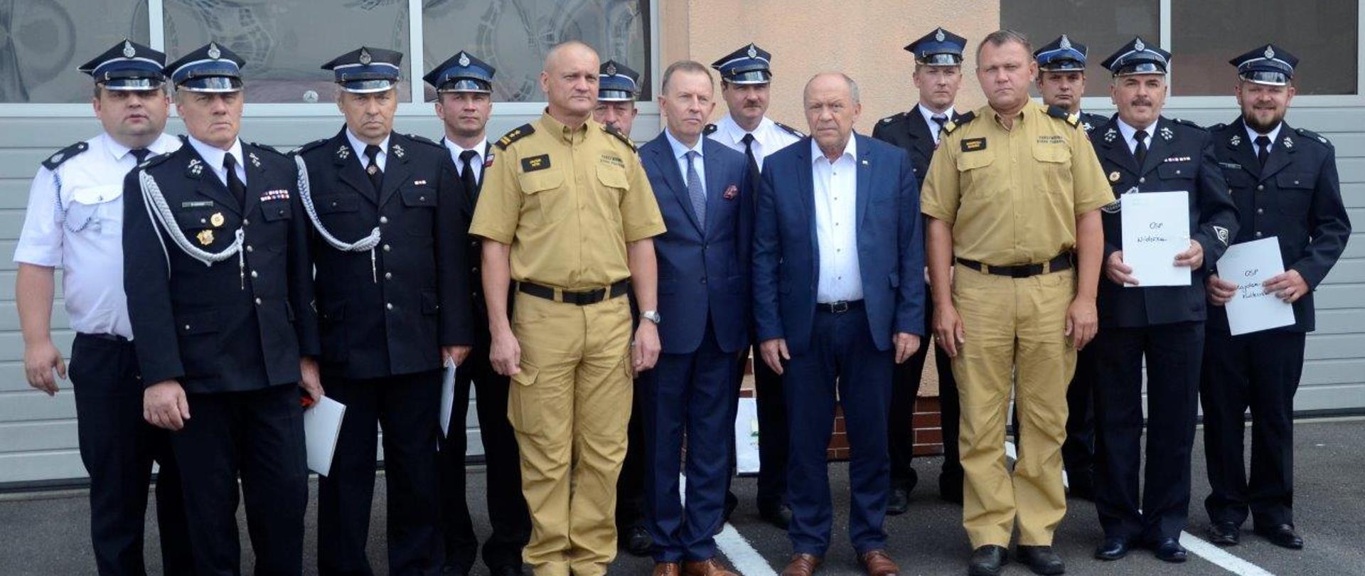 Przed budynkiem Jednostki Ratowniczo – Gaśniczej w Kolbuszowej na tle bram garażowych stoi grupa kilkunastu osób: dwóch strażaków PSP w mundurach w kolorze piaskowym, strażacy OSP w mundurach galowych oraz osoby cywilne w garniturach.