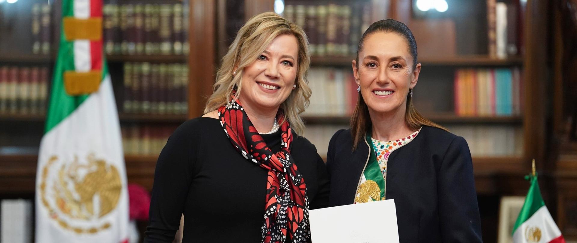 Na zdjęciu stoją obok siebie dwie uśmiechnięte kobiety w eleganckiej, reprezentacyjnej sali. Osoba po lewej to Ambasador RP w Meksyku, Agnieszka Frydrychowicz Tekieli, a po prawej to Prezydent Meksyku, Claudia Sheinbaum. Ubrane są odświętnie i każda trzyma w dłoniach białą teczkę z dokumentem — listem uwierzytelniającym. W tle widać regały z książkami oraz flagi Meksyku. Scena ma uroczysty, oficjalny charakter.