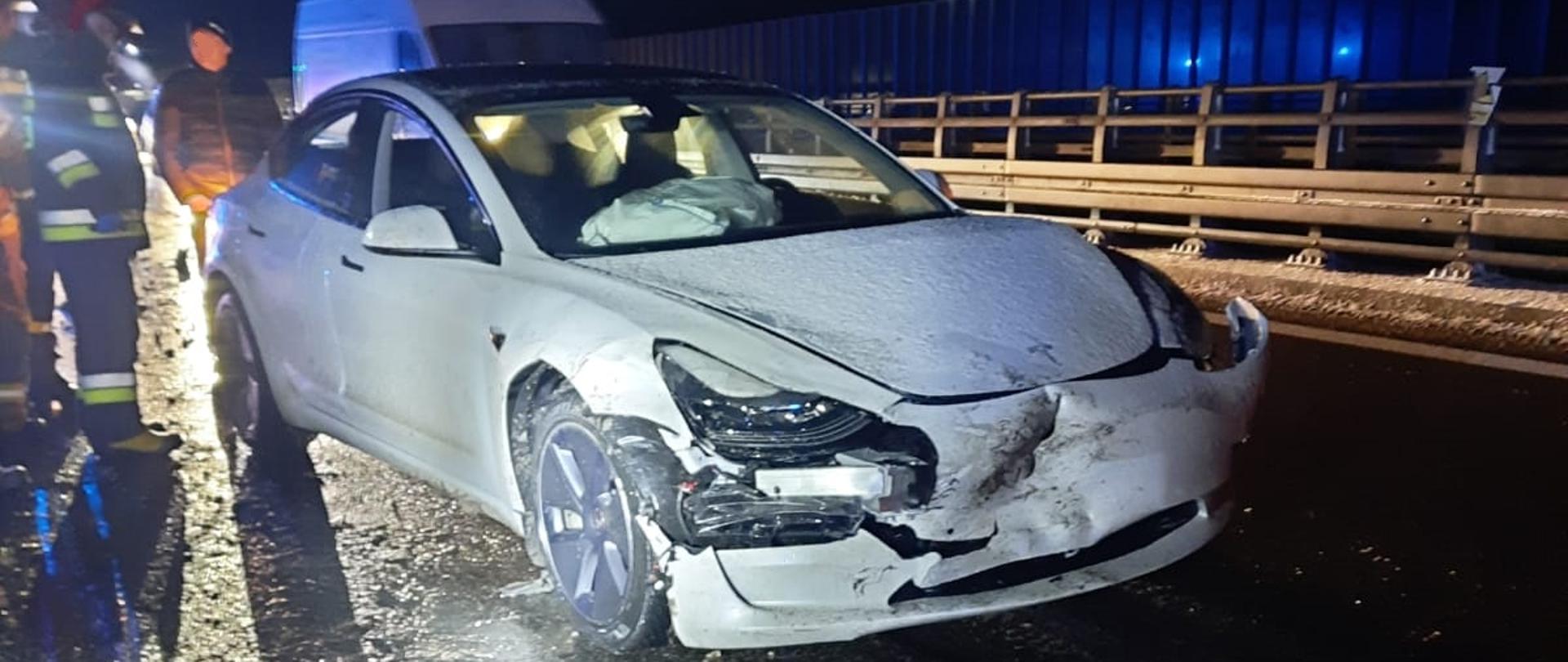 Zdjęcie obrazuje białysamochód elektryczny marki Tesla z uszkodzonym przodem. Obok pojazdu stoi właściciel oraz strażacy. W samochodzie wystrzeliły poduszki powietrzne. Na drodze widać oblodzenie, w tle widać niebieskie sygnały świetlne z samochodu pożarniczego.