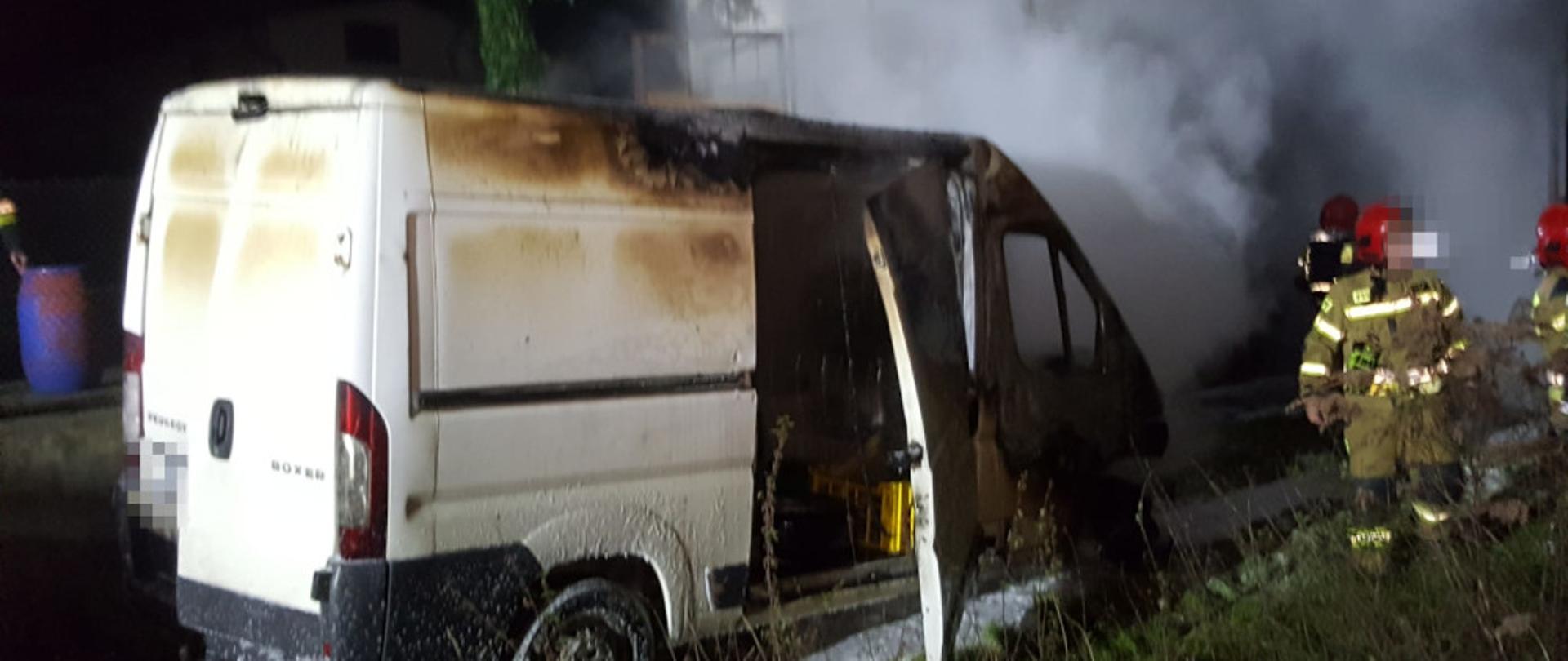 Pojazd dostawczy (biały peugeot) ze spalonym przodem oraz strażacy ubrani w ubrania specjalne i sprzęt ODO prowadzą działania polegające na ugaszeniu drewnianych elementów garażu.