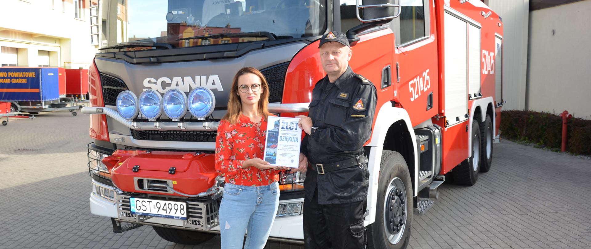 Zdjęcie przedstawia moment wręczenia podziękowań Komendantowi Powiatowemu, w tle ciężki samochód pożarniczy 
