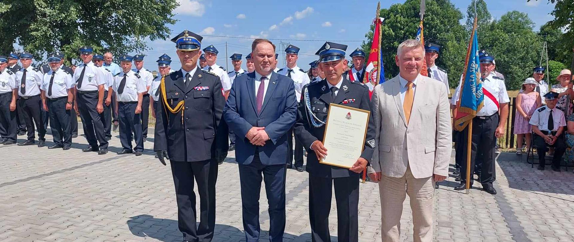 Zdjęcie przedstawia prezesa OSP Brzezie z aktem wręczenia jednostki OSP w struktury KSRG wraz z parlamentarzystami i zastępcą KW PSP w Kielcach. W tle pododdział towarzyszący
