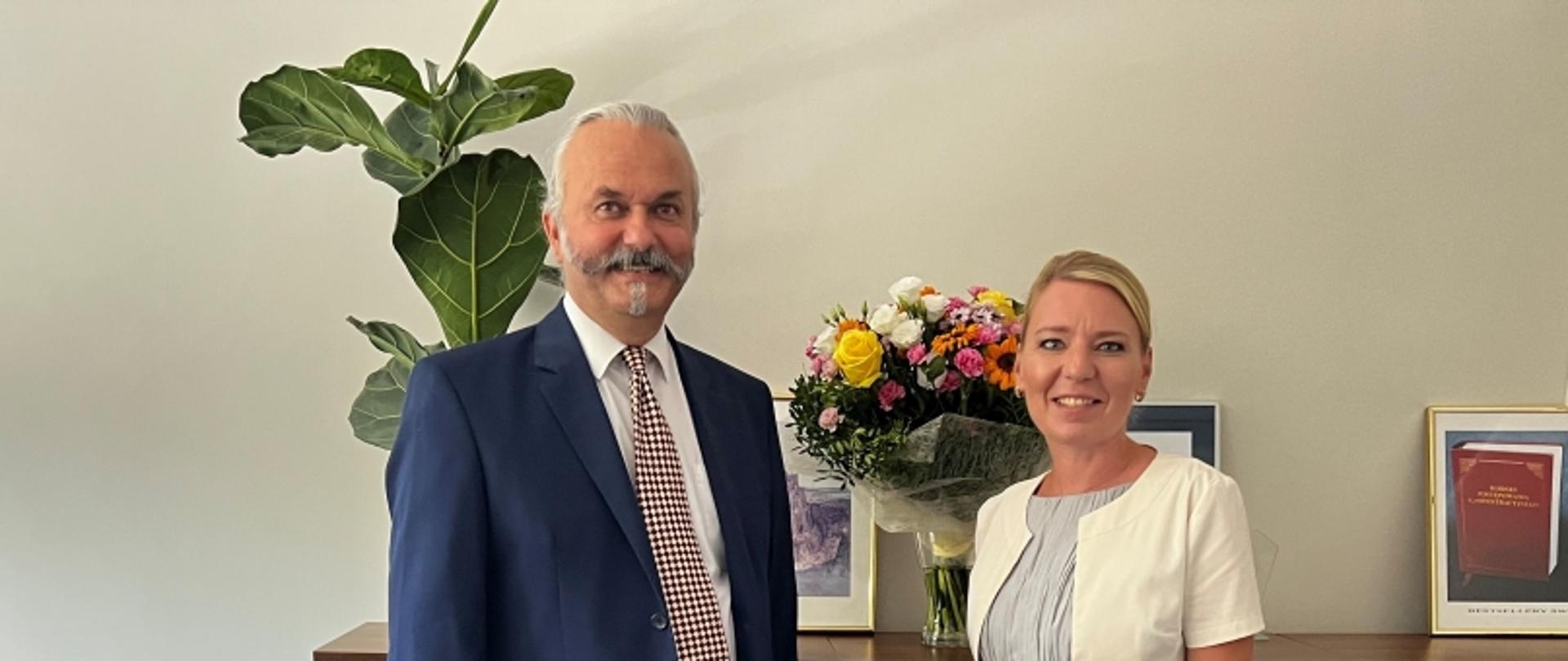 Dwie osoby na tle rośliny i stołu z kwiatami oraz oprawionymi dokumentami.