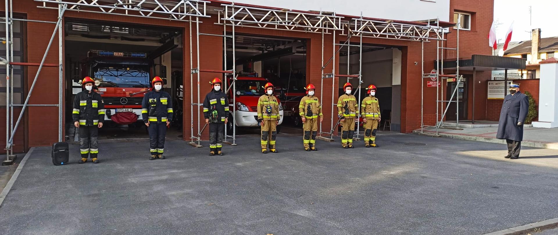 Przed budynkiem straży stoi na baczność w jednym rzędzie siedmiu strażaków. Za mini są trzy bramy garażowe otwarte przez które widać samochody strażackie. Po prawej stronie bokiem do strażaków stoi zastępca komendanta powiatowego ubrany w czarny płaszcz strażacki