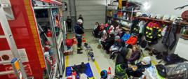 Dzieci siedzą i słuchają prelekcji strażaka. Za nim czerwony samochód ratowniczo gaśniczy