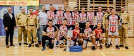 Na zdjęciu widoczni zawodnicy zwycięskiej drużyny z zawieszonymi na szyjach złotymi medalami w towarzystwie komendantów z rejonu województwa podkarpackiego w mundurach służbowych strażaka. Zawodnicy ubrani są w koszulki sportowe z napisem KM PSP w biało-czerwonych barwach. Jeden z zawodników znajdujący się w pierwszym rzędzie po prawej stronie zdjęcia trzyma puchar za 1 miejsce.
