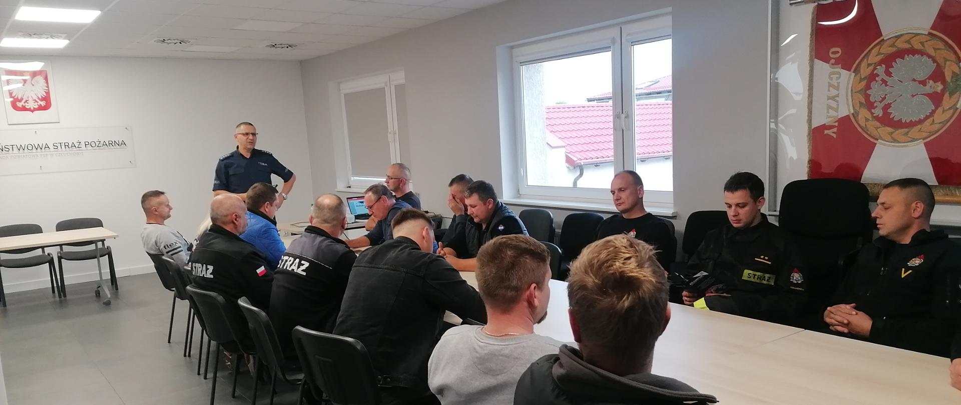 Zdjęcie przedstawia strażaków PSP i druhów OSP siedzących przy stole w sali Komendy Powiatowej Państwowej Straży Pożarnej w Człuchowie. Przy szczycie stołu stoi policjant prowadzący szkolenie z zakresu przepisów ruchu drogowego.