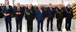 W rzędzie stoją wyróżnione osoby strażacy ubrani w mundury osoby cywilne w garnitury, na klapach mają przyczepione otrzymane medale. Z tyłu widać ustawionych strażaków również w mundurach galowych.