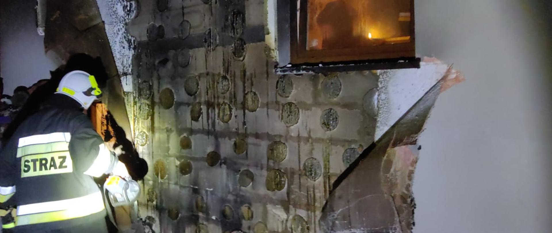 Zdjęcie przedstawia spaloną część elewacji na budynku jednorodzinnym. Pora nocna. Strażak ubraniu specjalnym świeci latarką na ugaszoną ścianę.