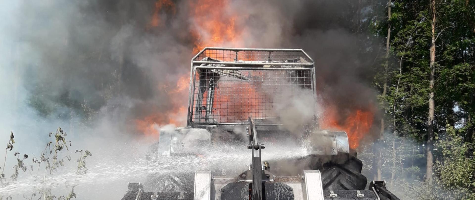 Na zdjęciu pokazano ciągnik leśny w ogniu stojący wśród lasu. Widać wysoki płomień i chmury dymu. 