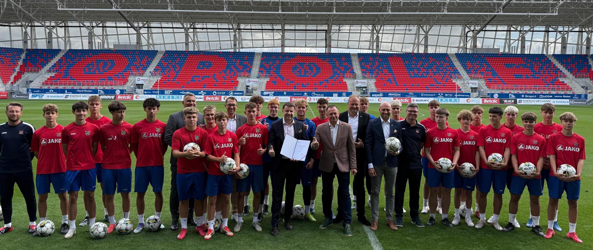 Zbiorowe zdjęcie przedstawiające wiceministra sportu i turystyki Piotra Borysa z członkami Akademii Odry Opole. Zdjęcie zrobione jest na murawie stadionu. W tle widać trybuny z napisem "Opole".