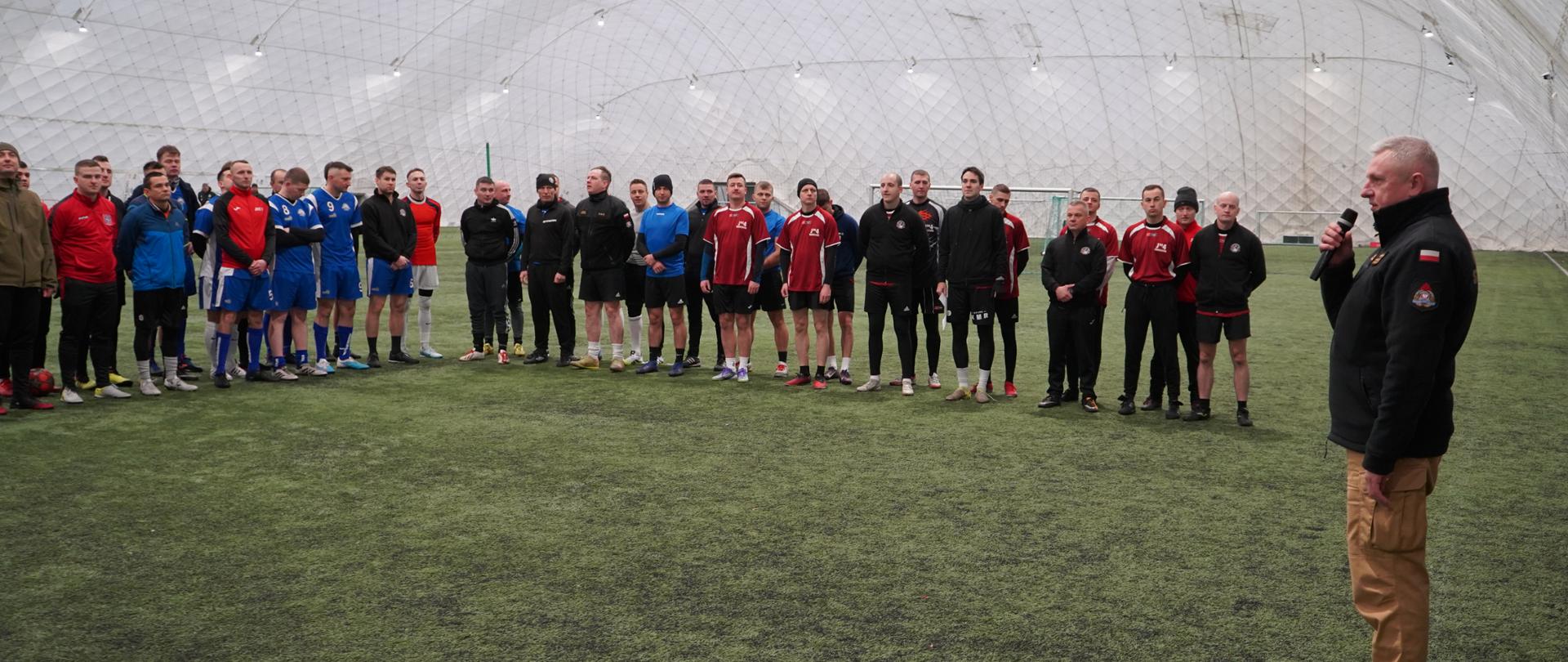 Zdjęcie przedstawia dużą grupę osób zgromadzoną wewnątrz zadaszonej hali sportowej o wysokim, białym, półprzezroczystym dachu w kształcie kopuły. Komendant Miejski PSP w trakcie przemówienia i powitania funkcjonariuszy na Turnieju