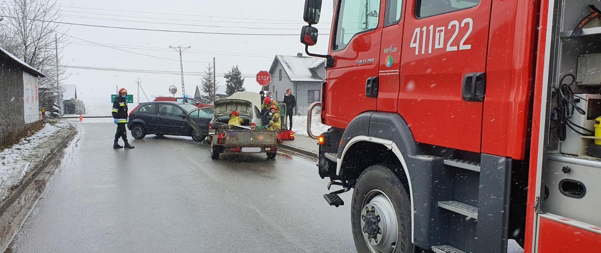 Zdjęcie przedstawia na pierwszym planie po prawej stronie lewą stronę pojazdu ratowniczo-gaśniczego GBA-Rt 3/16. W środkowej części zdjęcia widoczne są dwa uszkodzone samochody osobowe blokujące drogę powiatową. Do jednego z pojazdów przypięta jest lekka przyczepa. Przy pojazdach pracują strażacy w ubraniach specjalnych z założonymi czerwonymi hełmami na głowach. Zdjęcie zrobione w zimowej aurze przy opadach śniegu. 