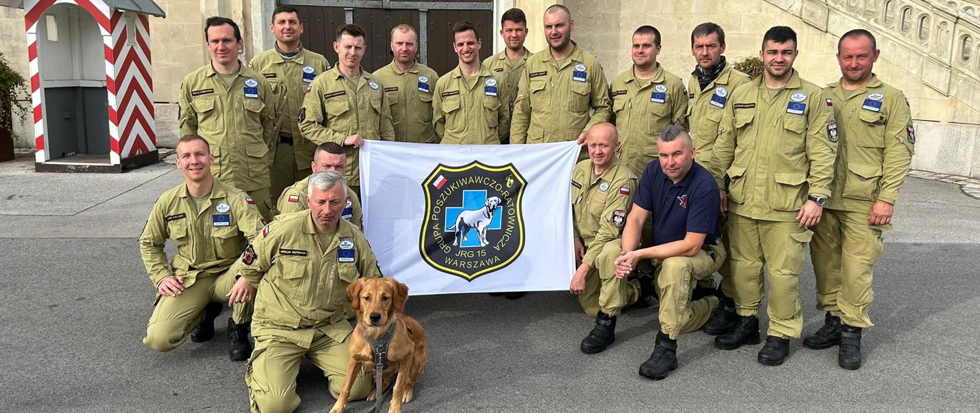 Strażacy z grupy specjalistycznej Poszukiwawczo-Ratowniczej wraz z psem. Na środku zdjęcia strażacy trzymają transparent z logo Grupy Poszukiwawczo-Ratowniczej.