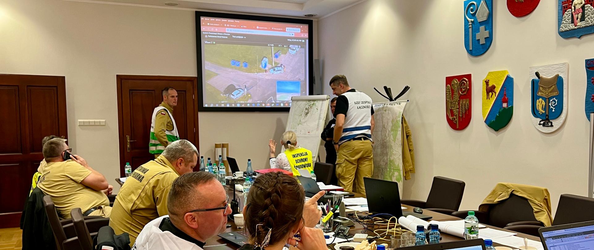 Strażacy Państwowej Straży Pożarnej przedstawiciele zakładu oraz instytucji siedzą za stołem na których rozłożone są laptopy na ekranie multimedialnym wyświetlany jest obraz z terenu działań.