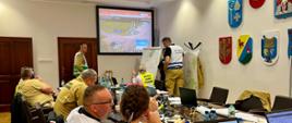 Strażacy Państwowej Straży Pożarnej przedstawiciele zakładu oraz instytucji siedzą za stołem na których rozłożone są laptopy na ekranie multimedialnym wyświetlany jest obraz z terenu działań.