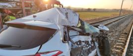 Widok uszkodzonego samochodu osobowego (z tyłu) znajdującego się na torach, obok strażacy i samochód gaśniczy.