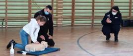 Zdjęcie przedstawia hale sportową na której znajduje się dziewczynka ucząca się prowadzić reanimację. Nadzoruje ją strażak a obok zdjęcie robi Pani policjant.