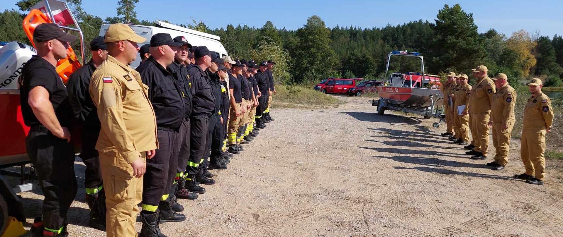 Ćwiczący strażacy stoją w dwuszeregu na zbiórce frontem do osób funkcyjnych.