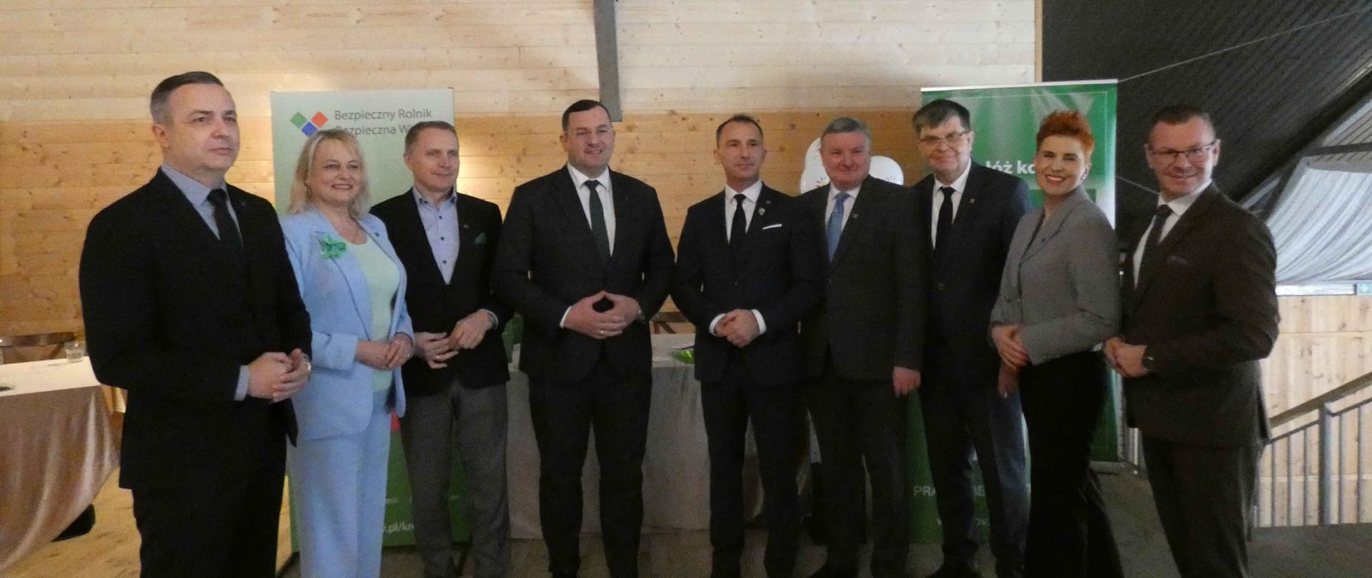 Dziewięć osób stoi obok siebie i pozuje do wspólnego zdjęcia w sali wydarzenia. Kobiety i mężczyźni ubrani w garnitury i eleganckie stroje stoją przed stoiskiem oraz roll-upami z materiałami informacyjnymi KRUS. W tle widoczne są drewniane ściany i elementy wystroju sali konferencyjnej. 