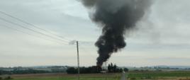 Na pierwszym planie widać z oddali czarne kłęby dymu unoszące się nad jednym z gospodarstw usytuowanym wśród pól. Zdjęcie wykonane z drogi asfaltowej prowadzącej do miejsca zdarzenia.