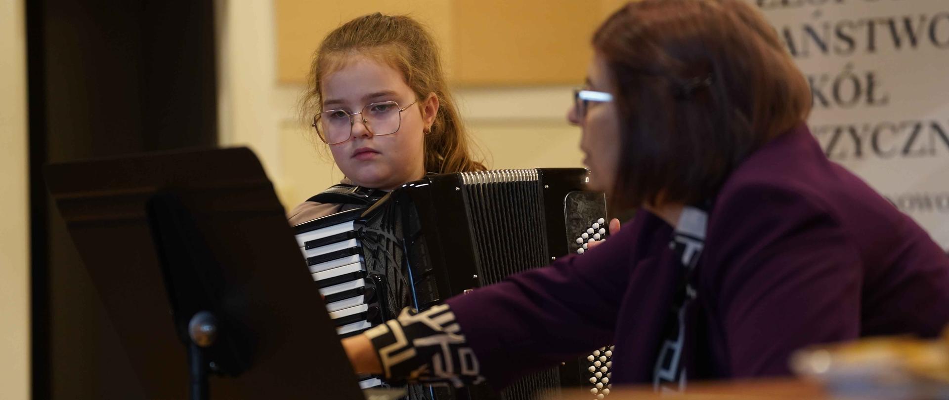zdjęcie przedstawia uczennicę młodszej klasy grającą na akordeonie, z boku prowadząca warsztaty udziela wskazówek, w tle baner szkoły