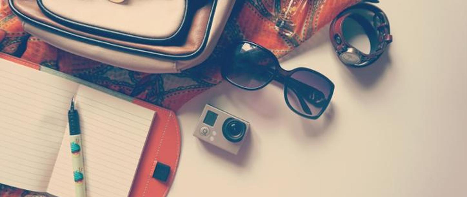 Na zdjęciu znajduje się materiał, na którym leży torebka oraz notatnik wraz z długopisem. Obok znajduje się aparat, okulary przeciwsłoneczne oraz zegarek. Powierzchnia, na której znajdują się wymienione przedmioty jest w odcieniach beżu.