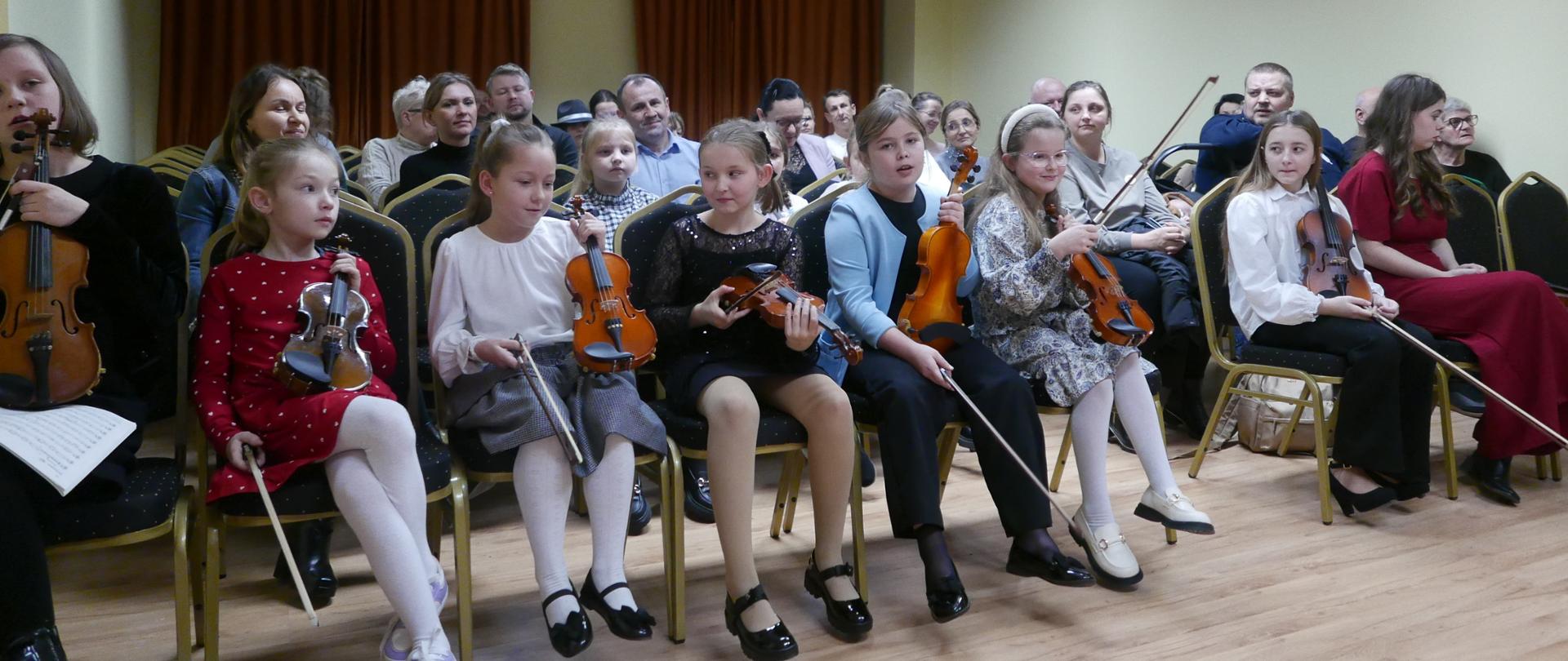 Klasa skrzypiec podczas koncertu. Widzimy dziewczynki siedzące w auli, trzymające skrzypce. Wszystkie za chwilę zagrają w ustalonej kolejności.