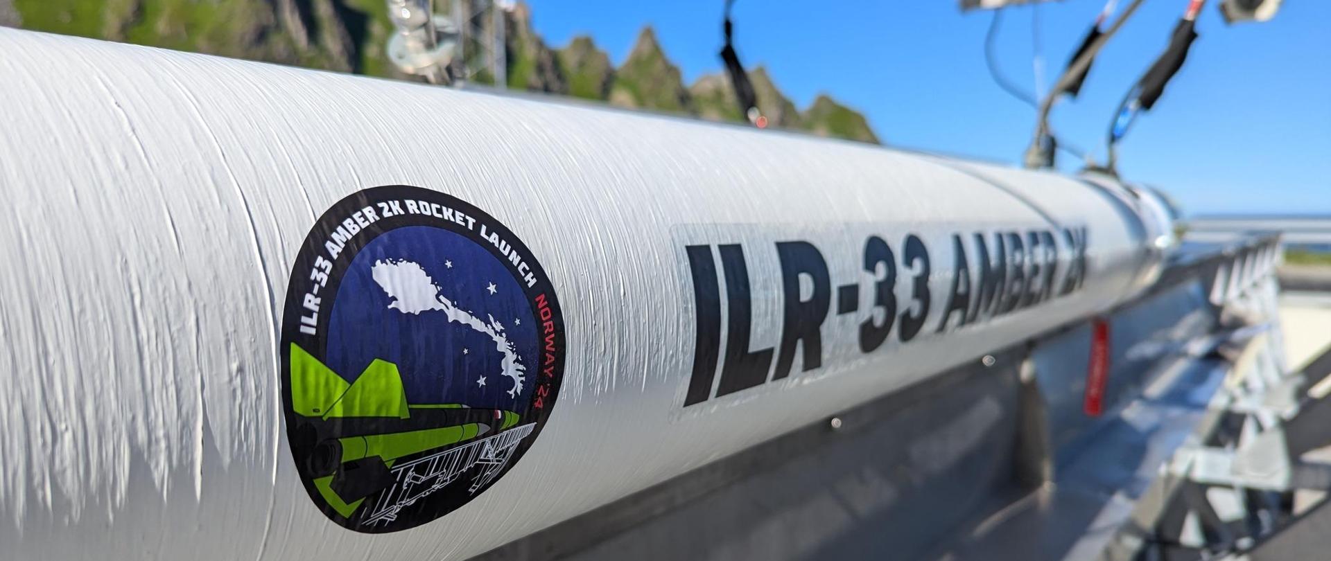 Zdjęcie na dworze, w słońcu, na poziomej platformie leży biały kadłub rakiety z napisem ILR-33 Amber, w tle zielone drzewa.